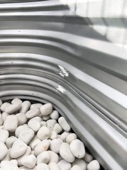 A close-up view of a corrugated metal window well with white decorative rocks at the base. The metal surface has minor scratches and wear, reflecting natural light. This functional design is commonly used for basement windows to allow light and ventilation while preventing soil erosion and water buildup.