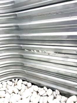 A close-up view of a corrugated metal window well with white decorative rocks at the base. The metal surface has minor scratches and wear, reflecting natural light. This functional design is commonly used for basement windows to allow light and ventilation while preventing soil erosion and water buildup.