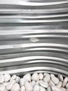 A close-up view of a corrugated metal window well with white decorative rocks at the base. The metal surface has minor scratches and wear, reflecting natural light. This functional design is commonly used for basement windows to allow light and ventilation while preventing soil erosion and water buildup.