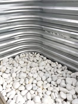 A close-up view of a corrugated metal window well with white decorative rocks at the base. The metal surface has minor scratches and wear, reflecting natural light. This functional design is commonly used for basement windows to allow light and ventilation while preventing soil erosion and water buildup.