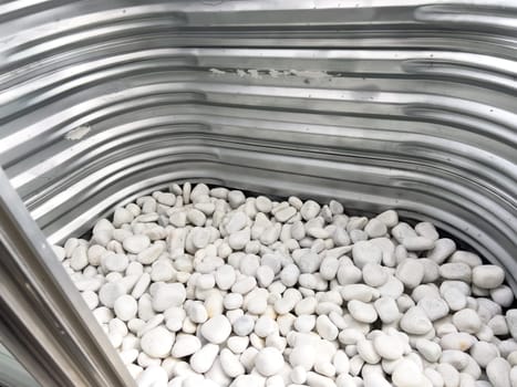 A close-up view of a corrugated metal window well with white decorative rocks at the base. The metal surface has minor scratches and wear, reflecting natural light. This functional design is commonly used for basement windows to allow light and ventilation while preventing soil erosion and water buildup.