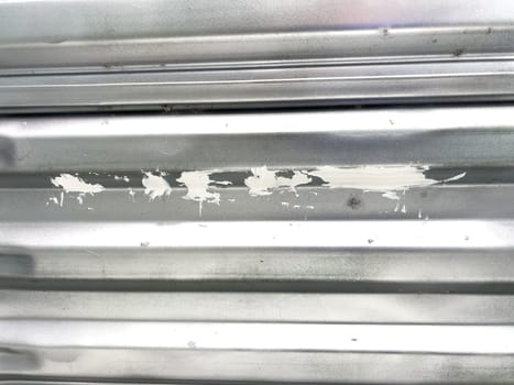 A close-up view of a corrugated metal window well with white decorative rocks at the base. The metal surface has minor scratches and wear, reflecting natural light. This functional design is commonly used for basement windows to allow light and ventilation while preventing soil erosion and water buildup.