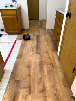 Broomfield, Colorado, USA-February 23, 2025-A kitchen undergoing flooring installation, featuring newly laid wood planks extending through the space. The underlayment is partially visible, with red tape securing it. Wooden cabinets line the kitchen, and installation tools are placed on the floor, indicating an ongoing renovation project.