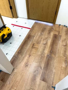 Broomfield, Colorado, USA-February 23, 2025-A kitchen undergoing flooring installation, featuring newly laid wood planks extending through the space. The underlayment is partially visible, with red tape securing it. Wooden cabinets line the kitchen, and installation tools are placed on the floor, indicating an ongoing renovation project.
