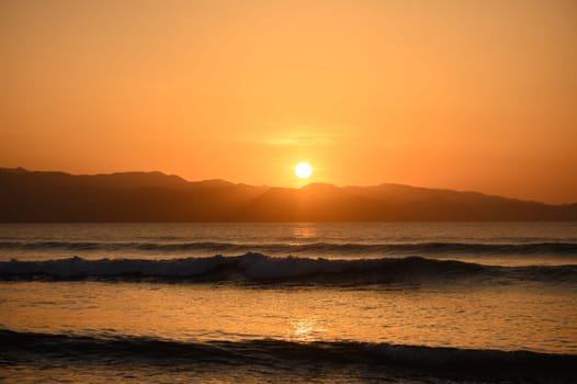 Warm hues stretch across the sky as waves gently lap against the shore at dusk, creating a tranquil atmosphere.