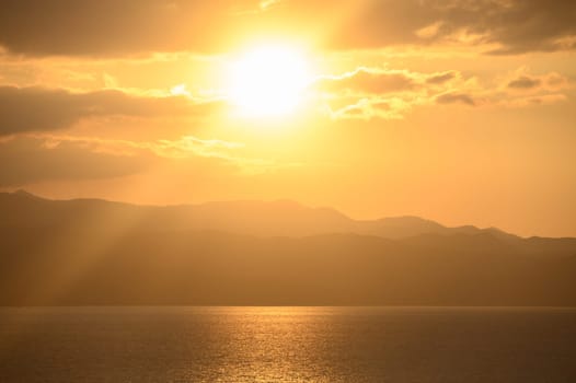 Warm sunlight bathes the horizon as clouds drift softly above serene waters and majestic mountains in the distance.