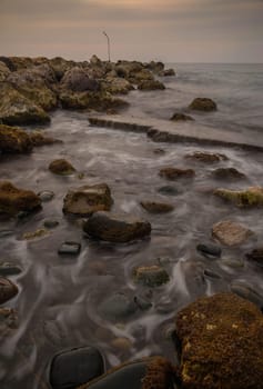 Dark clouds loom over the tranquil seashore as waves lap gently against rugged stones at twilight.