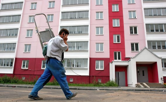 A worker with a ladder on his back talks on his phone while walking alongside a newly painted residential building, showcasing vibrant colors and windows.