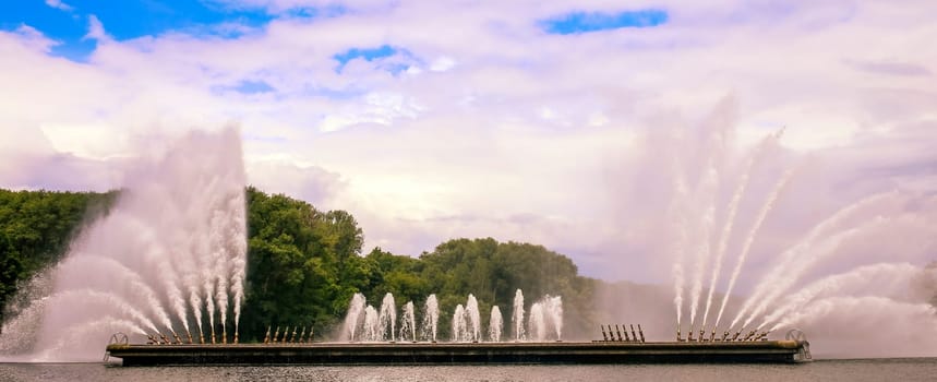Water jets create an enchanting display in a peaceful park, reflecting a blue sky and surrounded by lush greenery.