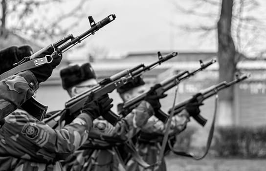 Men in military attire stand in formation, raising their rifles for a salute, showcasing discipline and tradition in a cold environment.