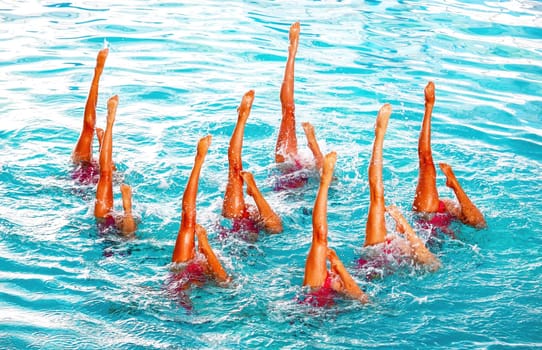 Synchronized swimmers showcase their teamwork and skill with raised legs in a pool under bright sunlight, creating beautiful patterns in the water.