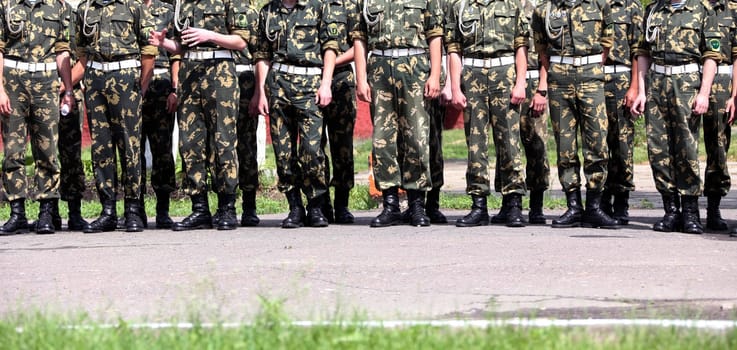 Groups of soldiers in camouflage uniforms stand at attention on a training ground under clear skies during summer.