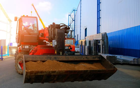 Construction workers are using heavy machinery to lift and transport soil in an industrial area filled with equipment and structures.