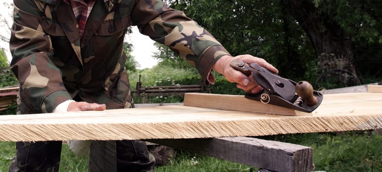 Craftsman works meticulously on a wooden plank using a hand planer in a peaceful outdoor workshop during daylight.