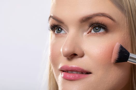 A professional makeup application showcasing a beautiful blonde woman gently applying blush with a brush