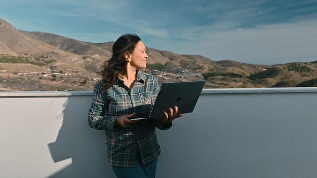 A woman standing on a balcony using a laptop, enjoying the scenic mountain landscape. The setting captures the essence of remote work lifestyle, blending productivity and nature.