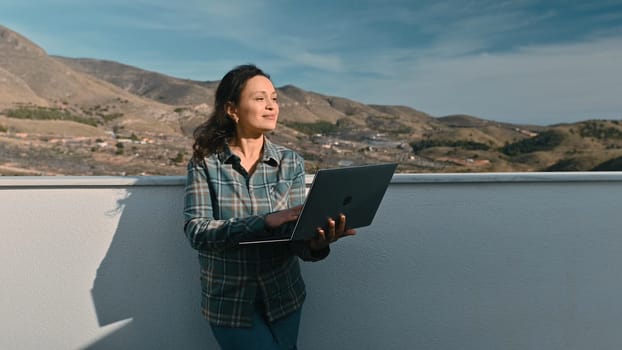 A woman stands outdoors holding a laptop, embracing remote work amidst a picturesque mountain landscape, blending productivity with relaxation.