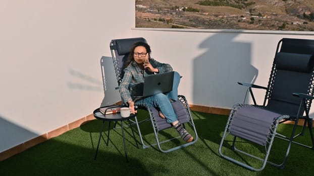 A woman working remotely on a laptop with a phone call on a sunny outdoor space.