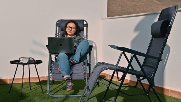 A woman sits comfortably on a reclining chair outdoors, using her laptop for remote work. Surrounded by sunlight and a serene setting, she enjoys the flexibility of a digital nomad lifestyle.