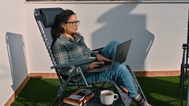 A woman sits in a reclining chair on a sunny terrace, using a laptop. She represents the concept of remote work and enjoying an outdoor office, showcasing flexibility in work environments.