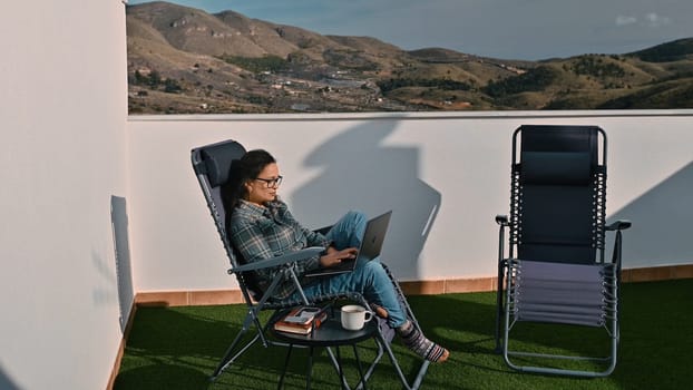 A woman enjoys working remotely on a laptop while lounging on a terrace overlooking stunning mountains. A second empty chair and table with items add a cozy ambiance to the space.