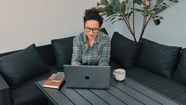 A woman focuses on her laptop while sitting outside on a cozy patio couch with a cup of coffee, surrounded by a relaxed atmosphere and greenery, exemplifying remote and outdoor work lifestyle.