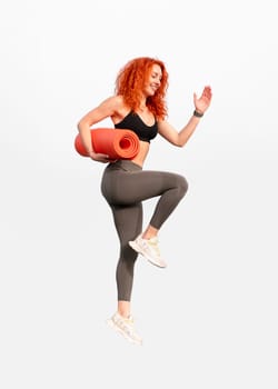 Happy jogger woman holding rolled up exercise mat in midair against gray background. Young female in sportswear is smiling while jumping in studio
