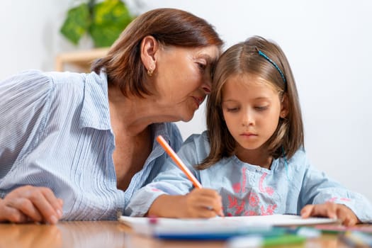 Loving grandmother whispers secret into granddaughter's ear as she colors in book at home. The picture captures warm, loving moment filled with affection and artistic activity