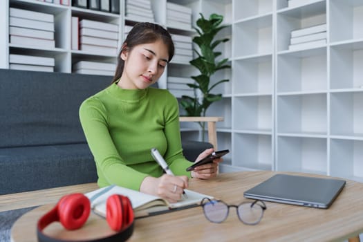 Woman sitting at a table with a laptop and cellphone using smartphone to take notes and work in a cozy home.