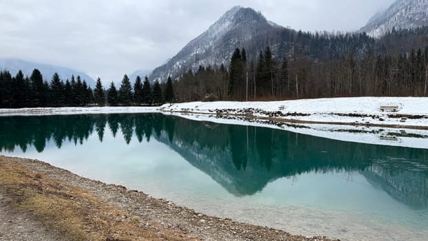 Emerald Winter Lake with Alpine Mountain Reflections. A stunning winter landscape featuring a crystal-clear emerald lake reflecting the surrounding snow-covered mountains and evergreen trees. The calm water creates a perfect mirror image, enhancing the serene and tranquil atmosphere. Snow-dusted benches along the shore invite peaceful contemplation, making this a picturesque and relaxing nature scene.