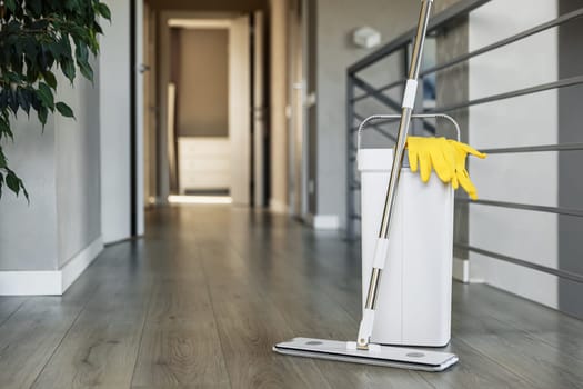 Cleaning supplies: mop, gloves, bucket with wringer sitting on wooden laminate floor in modern living room, wet equipment tools, housekeeping concept. Emphasizing home sanitation, cleanliness, hygiene