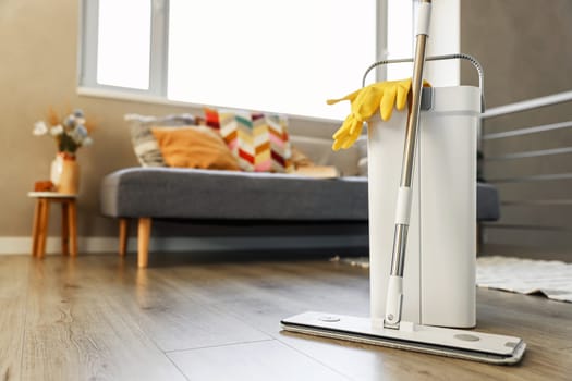Cleaning supplies: mop, gloves, bucket with wringer sitting on wooden laminate floor in modern living room, wet equipment tools, housekeeping concept. Emphasizing home sanitation, cleanliness, hygiene