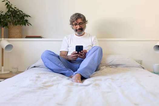 Mature man using phone relaxing on bed at home. Lifestyle concept.