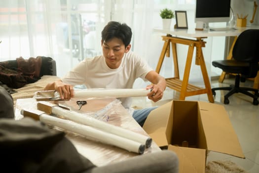 Young asian man unpacking a table leg preparing to assemble a new piece of furniture in his living room.
