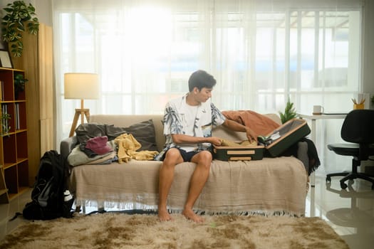 Cheerful young man in a tropical shirt packing his suitcase for summer trip.