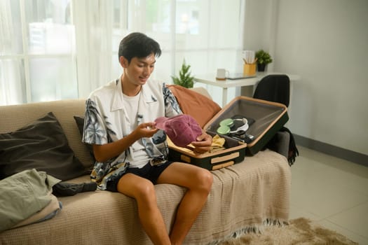 Cheerful young man in a tropical shirt packing his suitcase for summer trip.