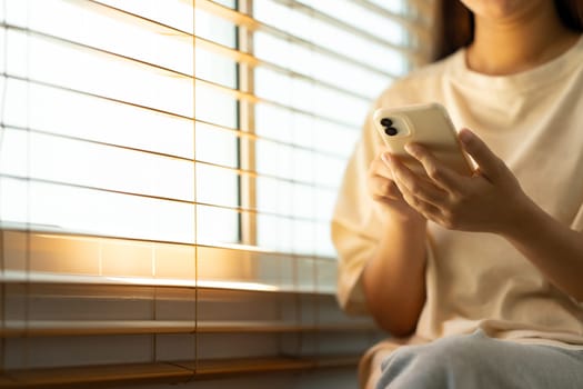 Young woman sitting near a sunlit window and using smartphone.