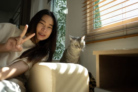 Cheerful asian woman posing with her adorable gray cat near sunlit window in a living room.