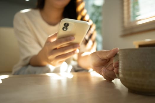 Young woman holding a cup of coffee and using mobile phone in bright living room.