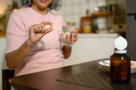 Smiling senior woman holding a fish oil capsule and a glass of water sitting in a cozy kitchen.