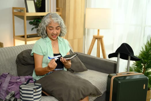 Excited senior woman holding a miniature airplane and passport sitting on couch with travel accessories.