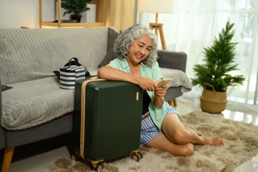 Cheerful mature woman sitting on the floor next to her suitcase and using smartphone.