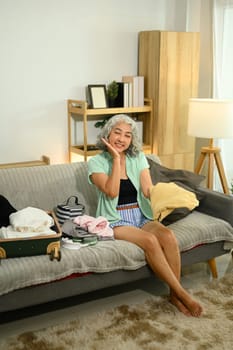 Happy senior woman is sitting on a cozy couch, preparing luggage for a summer trip.