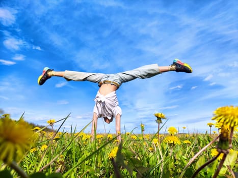 A person performs a handstand in a vibrant field of dandelions, enjoying the sunny weather and clear blue sky above.