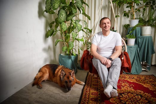 A man sits comfortably in a swinging chair while his German Shepherd lies nearby amid lush greenery.