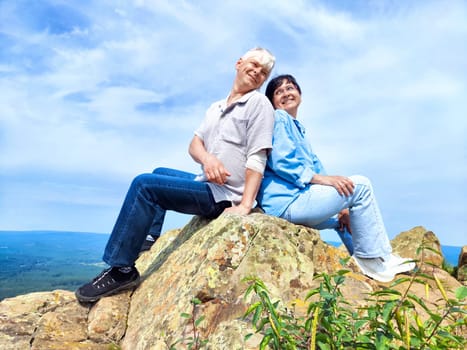 Two people sit back to back on a large rock, smiling and enjoying the beautiful view of the landscape below.