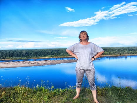 A middle-aged female blogger smiles while taking a selfie against a stunning river landscape in summer.