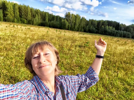 Mature blonde travel blogger takes a cheerful selfie in a vibrant field surrounded by trees on a sunny day.