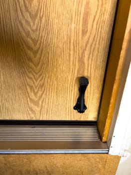 Close-up of a wooden door featuring a black metal kickstand near the bottom. The door is set within a metal threshold, and the surrounding floor has a textured surface. The kickstand is used to keep the door propped open, providing convenience and functionality.