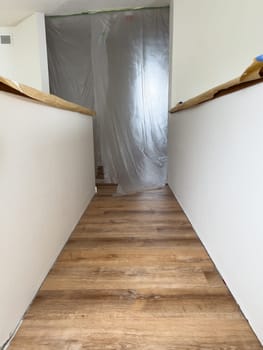 A freshly renovated upstairs hallway featuring new wood-look flooring. The walls are painted white, and protective plastic sheeting covers an entryway, indicating ongoing construction or remodeling. Handrails are partially wrapped for protection during the renovation process.
